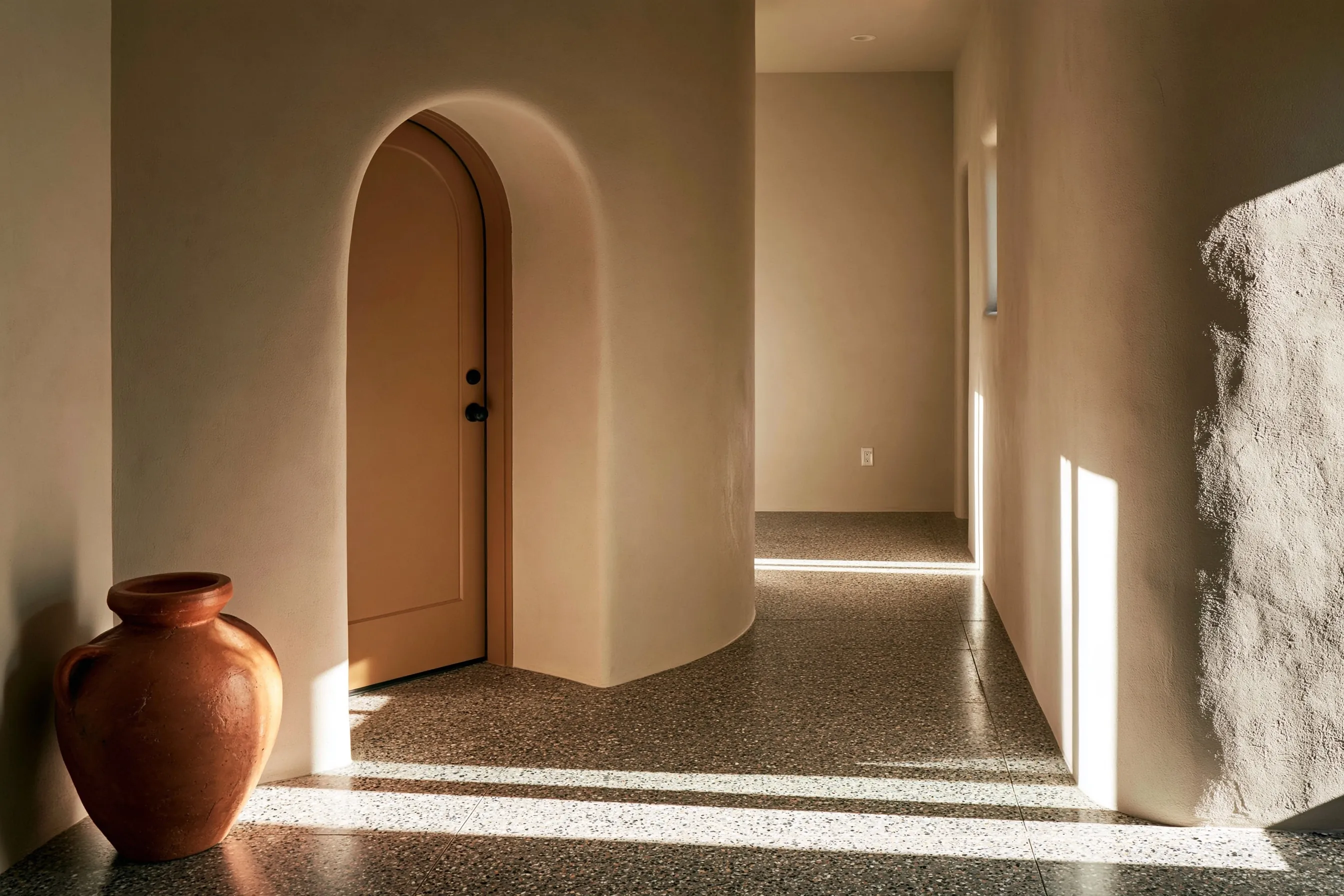 Entry hall and corridor with a salt-and-pepper fine-aggregate polished concrete floor, an arched stucco doorway and a terracotta pot in daylight
